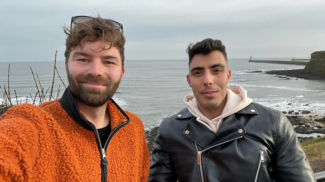 This picture is a selfie between two men in front of the sea. The man holding the phone is dressed in an orange sweater with glasses on top of his head. The man on the right is wearing a leather jacket.